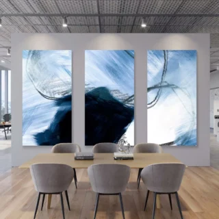 A contemporary office space features a wooden dining table surrounded by chairs. Behind the table, three large acoustic canvas's in shades of blue and white add a touch of modern art to the space.