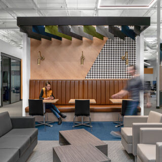 A modern office interior with a suspended Ezobord ceiling baffle installation. The baffles are arranged in a geometric pattern and feature a mix of colors and materials. A woman is sitting at a table in the foreground, and a man is walking in the background. The space also includes comfortable seating areas with sofas and chairs.
